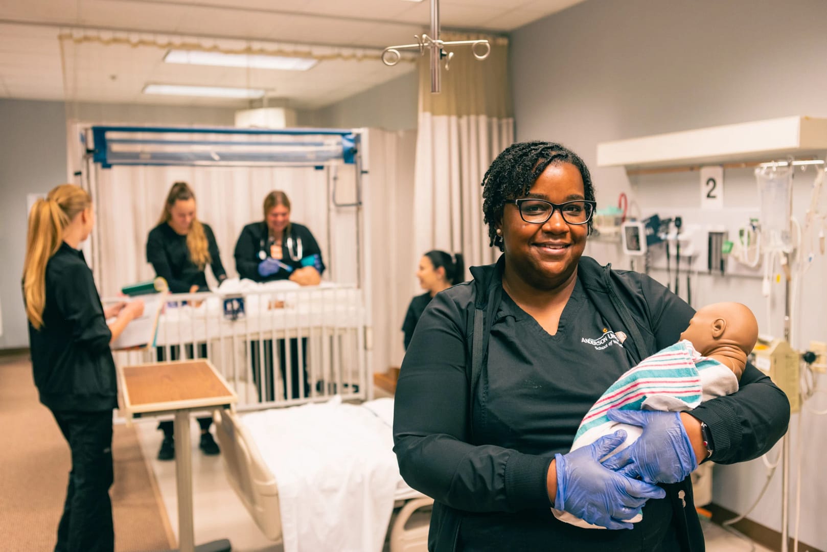 absn student smiles in the foreground while holding a baby mannequin and other nursing exercise clinical skills in the background