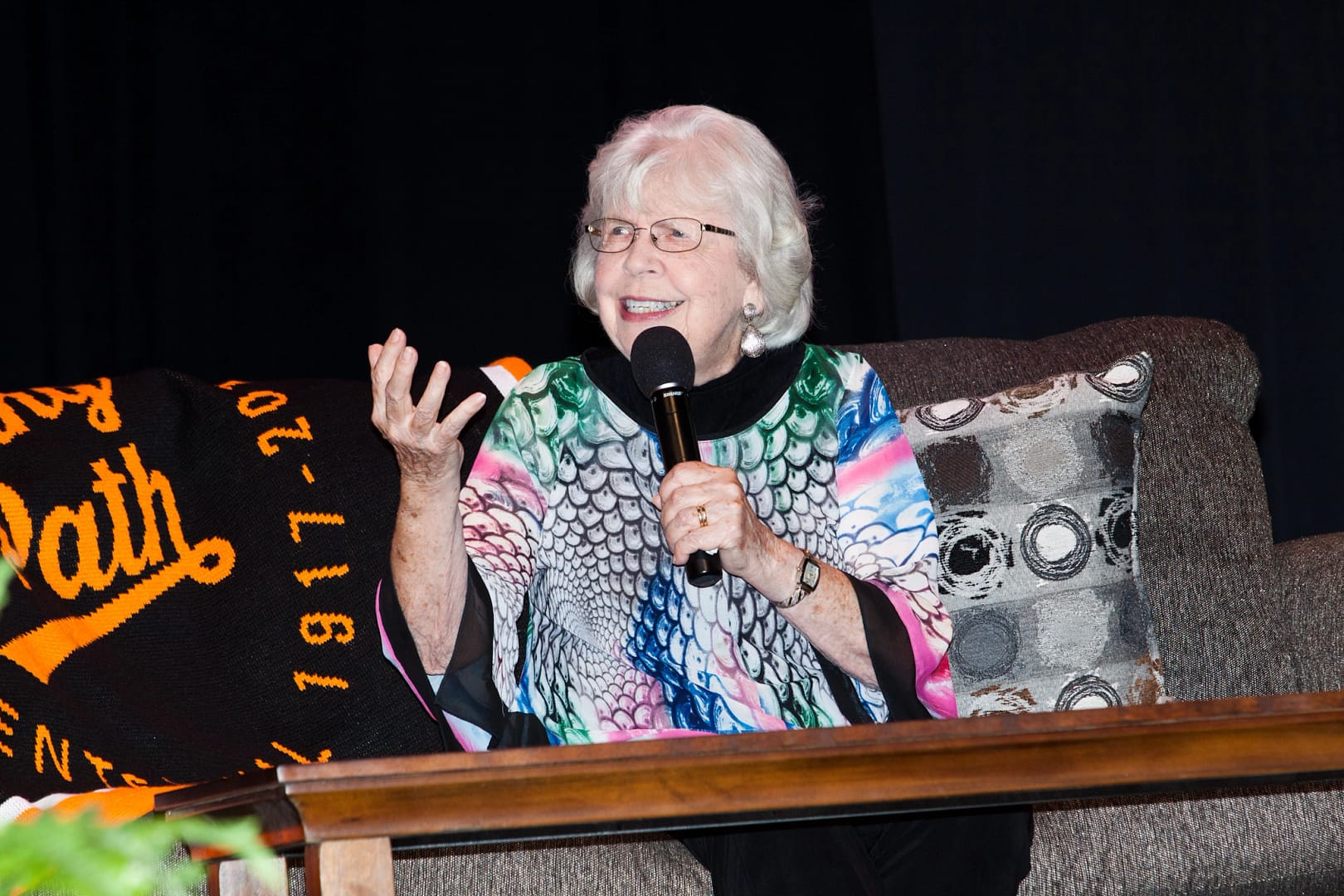 Ann Smith sits on a couch while speaking with a microphone in her hand